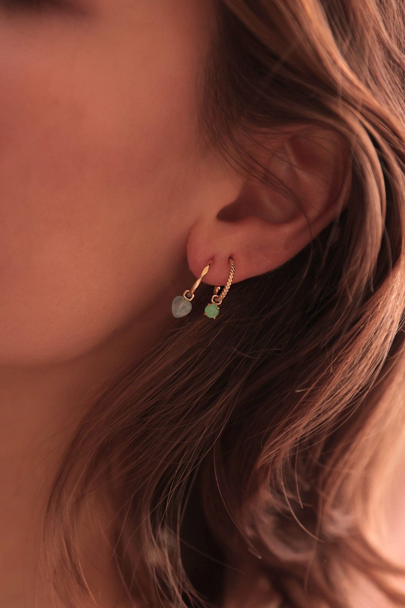 Kvinde med øreringe i ægte guld fra carré jewellery med en hjerteformet ørering med blå sten og en charm til hoops i grøn chrysopras